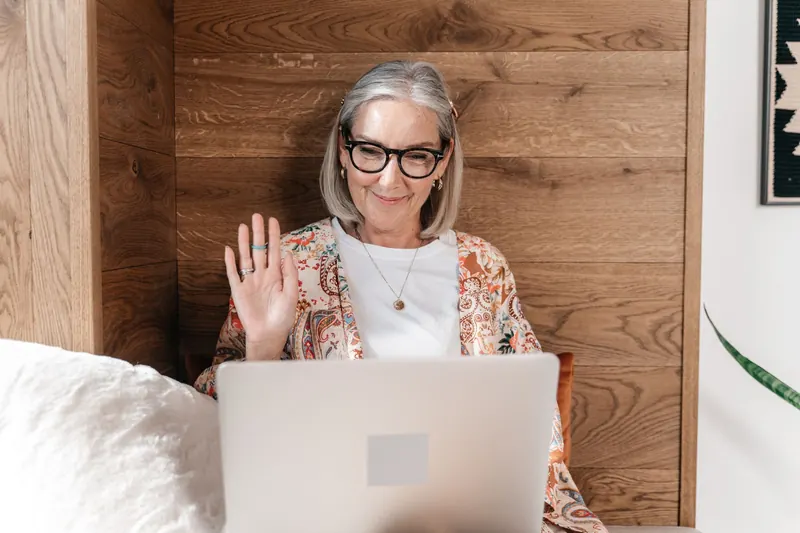 A woman with gray hair waving during a video call on her laptop