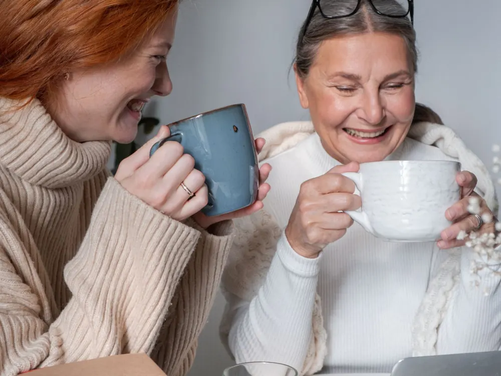 An older mother and her adult daughter laughing together over cups of coffee