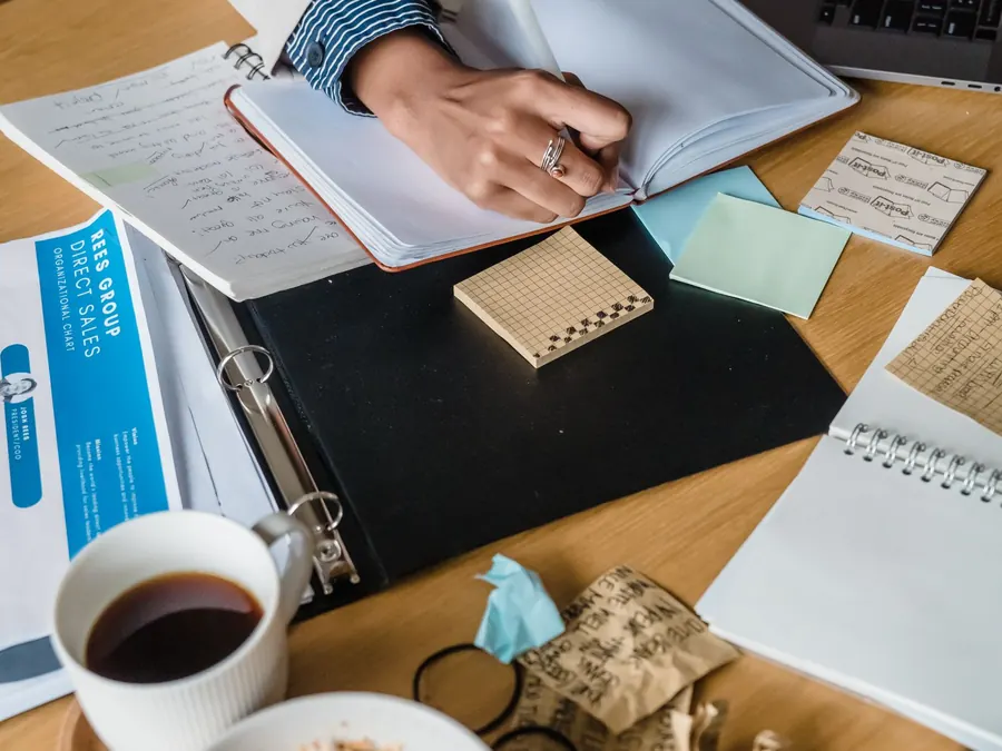 Hands writing in a notebook next to a cup of coffee and a folder of papers